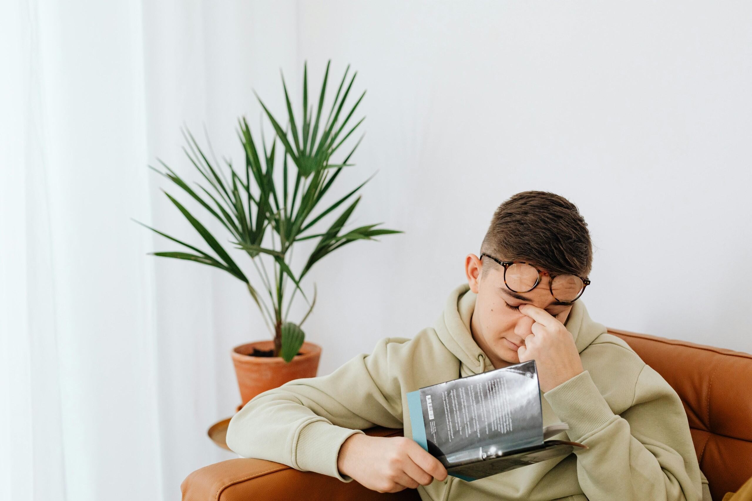 a man rubs his eyes while sitting on a couch and holding a book