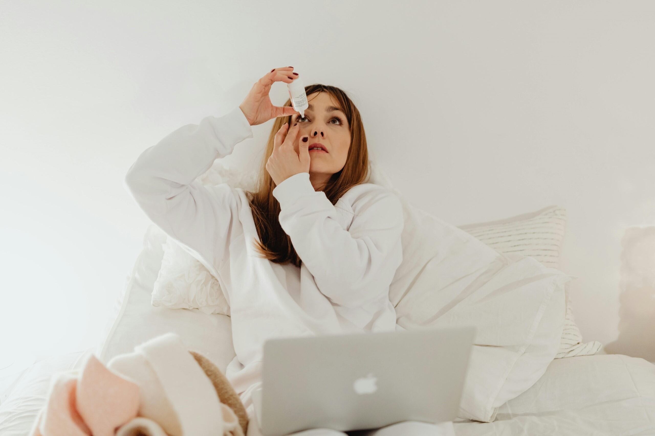 a woman puts eye drops in one eye while sitting on a couch